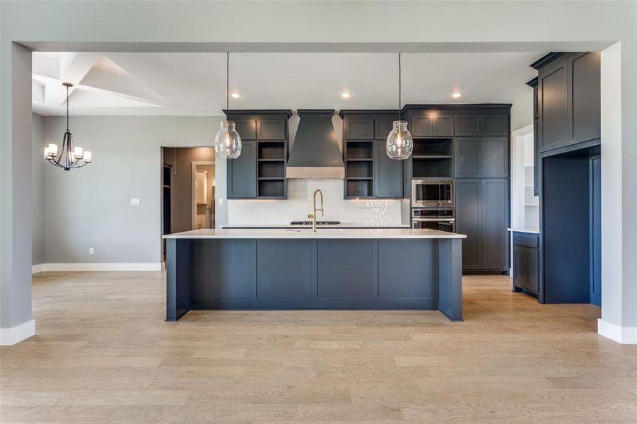 Kitchen with hanging light fixtures, a kitchen island with sink, custom range hood, backsplash, and light wood-style flooring Kitchen with hanging light fixtures, a kitchen island with sink, custom range hood, backsplash, and light wood-style flooring