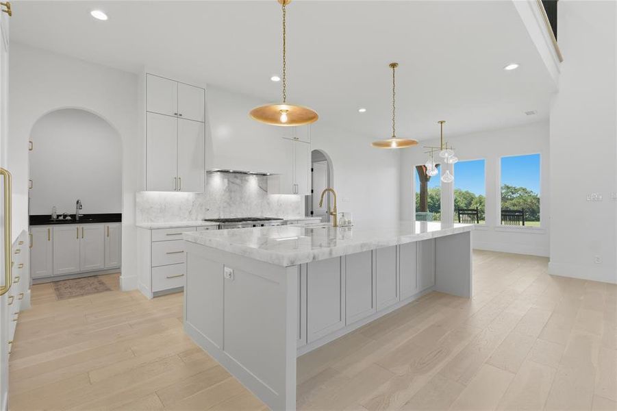 Gourmet kitchen with a large central island, light wood-finish flooring, white cabinetry, marble countertops, and gold-toned pendant lighting fixtures