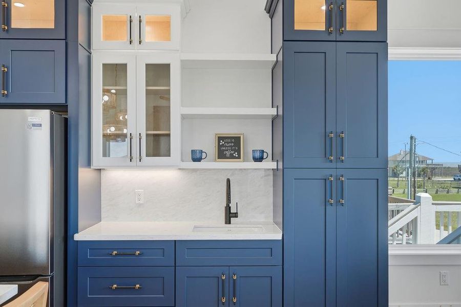 This kitchen features elegant blue cabinetry with gold hardware, white countertops, and a built-in sink. It includes glass-front upper cabinets, open shelving, and a view of the outdoors through a nearby window, creating a bright and stylish space.