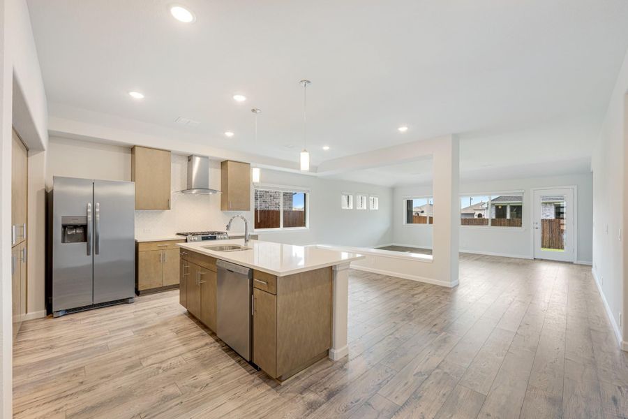 Open kitchen with center island, wood cabinets, stainless appliances, and light hardwood floors opening to living area