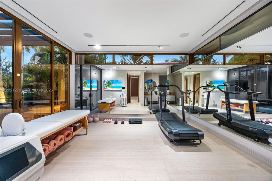 Full private gym and wellness suite with treadmills, kettlebells, yoga mats, massage table, full-wall mirror, wall-mounted TV, light wood floors, clerestory windows, floor-to-ceiling bronze-framed glass walls, and adjacent treatment or steam room.