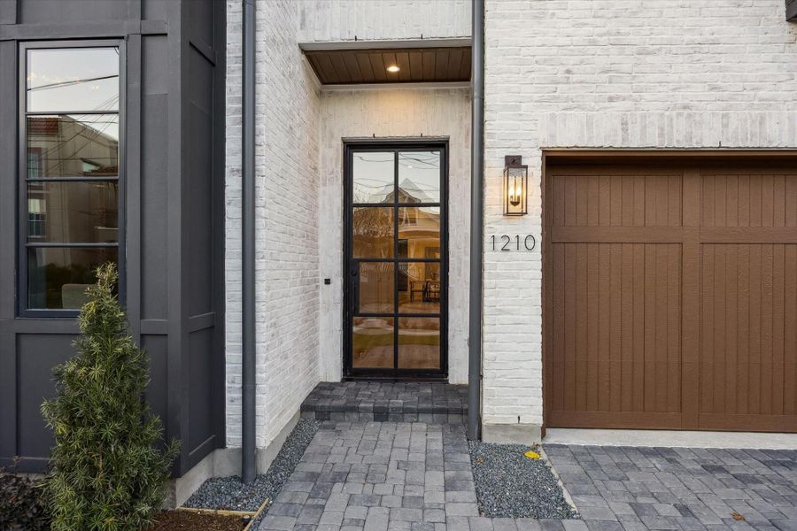 The front entrance features brick, pavers, and contrasting garage door and architectural elements. Pavers line the path and a dramatic obsidian steel pivot door previews the splendor to come!