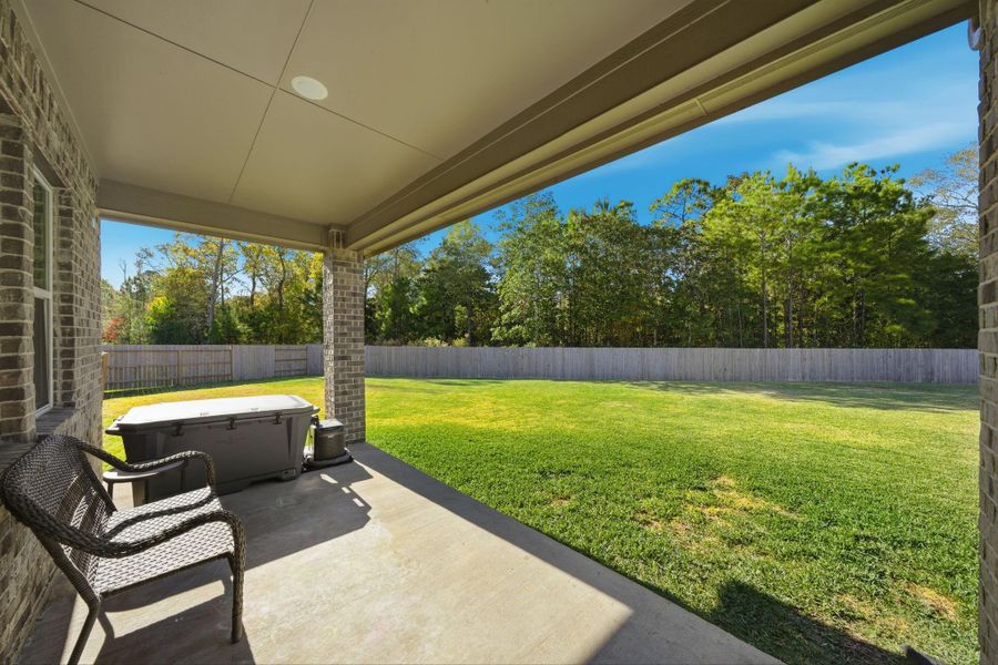 This photo shows a covered patio with brick accents overlooking a spacious, fenced backyard. Ideal for outdoor relaxation and entertainment, it offers a peaceful view of lush greenery.