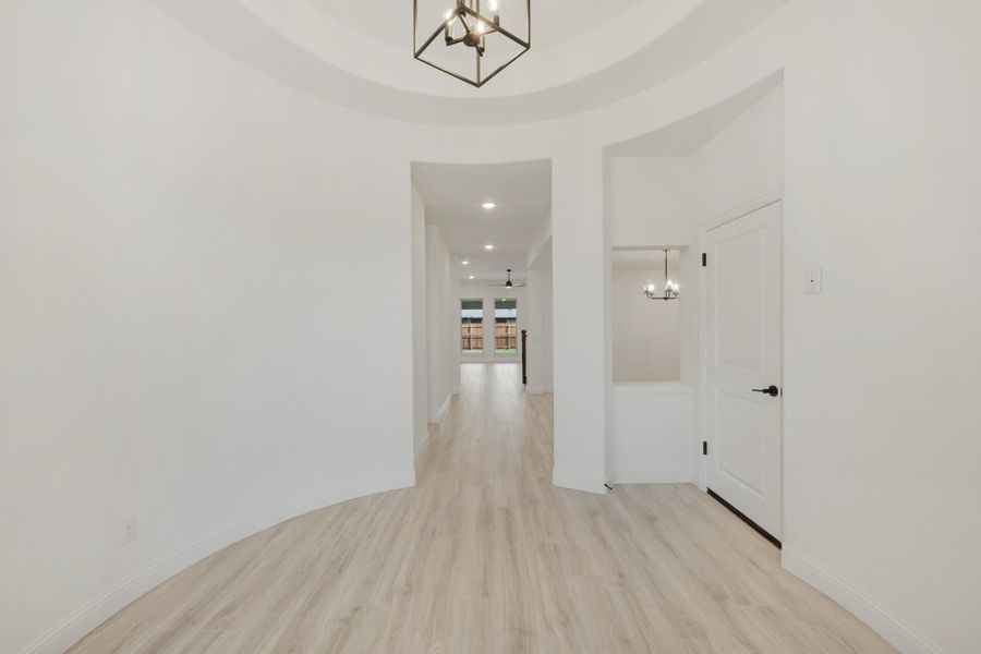 Curved entryway with tray ceiling, geometric chandelier, and light wood floors leading to main living area