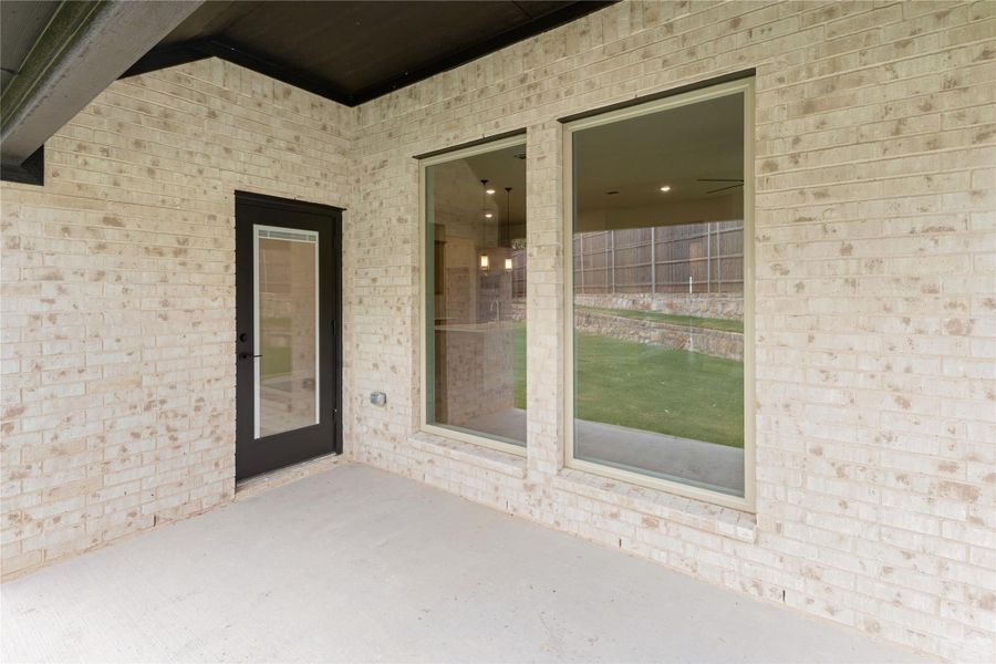 Covered concrete patio with brick exterior wall, black glass door, and large windows overlooking backyard Covered concrete patio with brick exterior wall, black glass door, and large windows overlooking backyard