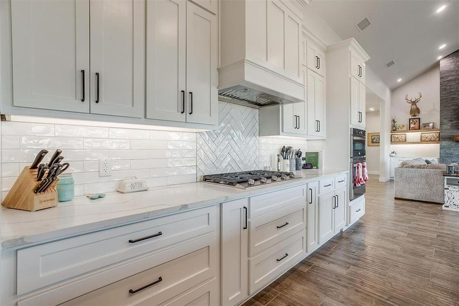 Kitchen featuring vaulted ceiling, white cabinetry, decorative backsplash, custom exhaust hood, and light stone countertops