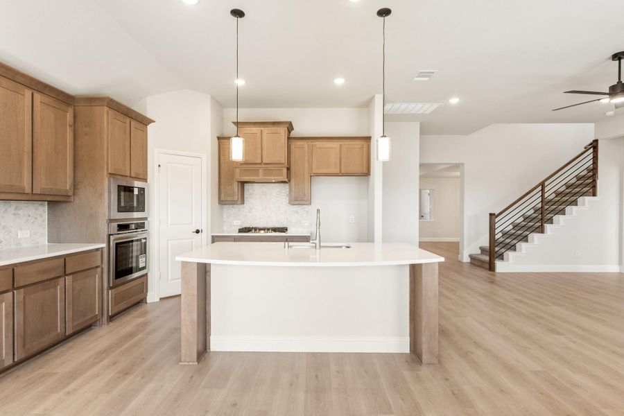 Kitchen with wood cabinets, large center island, stainless appliances, and light hardwood floors open to living area with staircase.