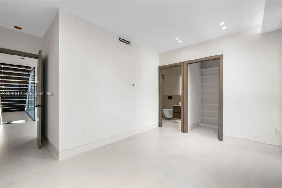 Neutral interior space featuring large-format tile flooring, recessed ceiling lighting, and wood-trim doorways leading to a built-in shelving unit and a bathroom Neutral interior space featuring large-format tile flooring, recessed ceiling lighting, and wood-trim doorways leading to a built-in shelving unit and a bathroom