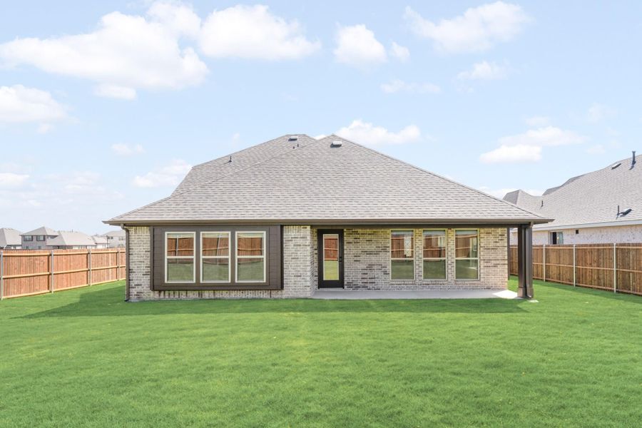 Rear view of single-story brick home with covered patio, large windows, and fenced backyard with green lawn.