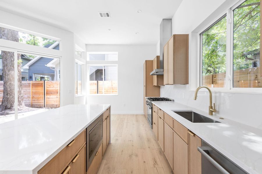 Kitchen with light wood-type flooring, light countertops, stainless steel appliances, modern cabinets, and wall chimney exhaust hood Kitchen with light wood-type flooring, light countertops, stainless steel appliances, modern cabinets, and wall chimney exhaust hood