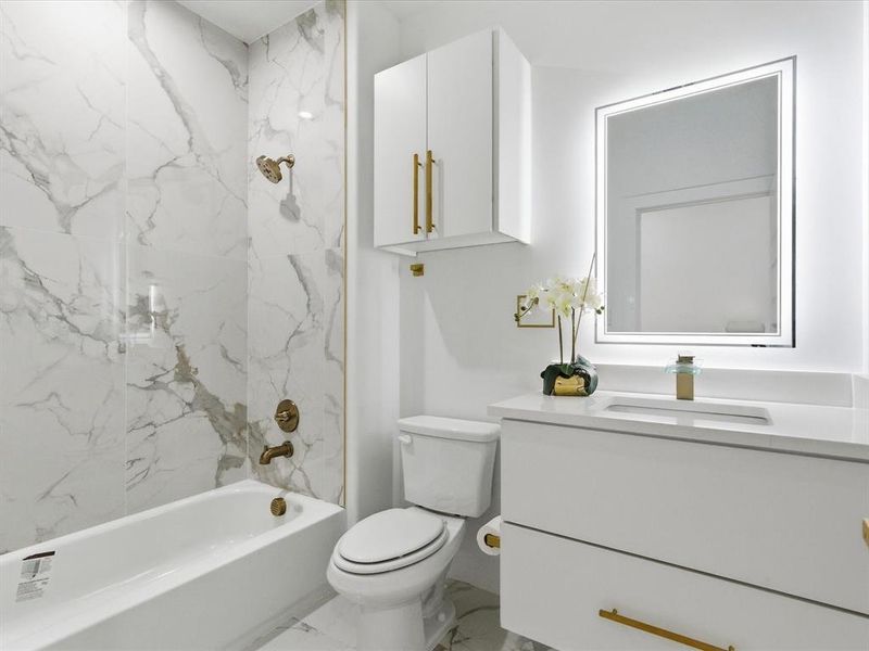 Brightly lit bathroom featuring a white vanity with gold-tone hardware, an illuminated mirror, a white toilet, a white bathtub, and large format wall tile