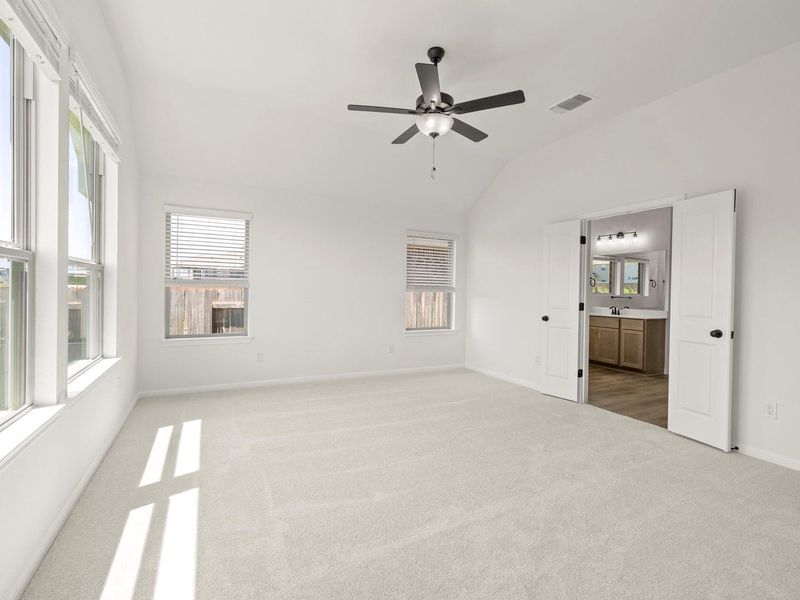 Spacious room featuring vaulted ceilings, light-colored carpeting, multiple windows with blinds, a ceiling fan with integrated lighting, and double doors opening to a bathroom with wood-finish flooring and a dual vanity
