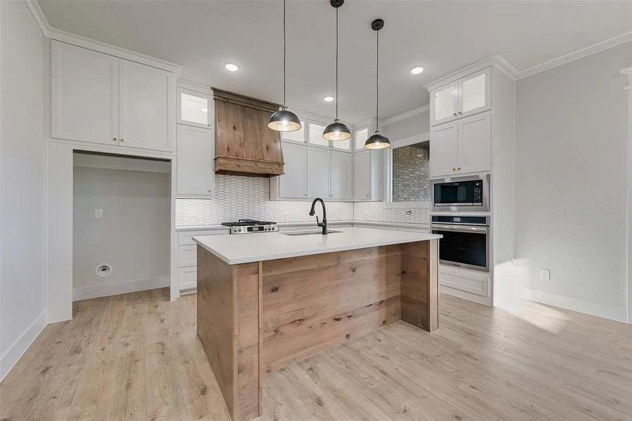 Two tone kitchen featuring glass fronted cabinets, a center island with sink, stainless steel appliances, hanging light fixtures, and crown molding