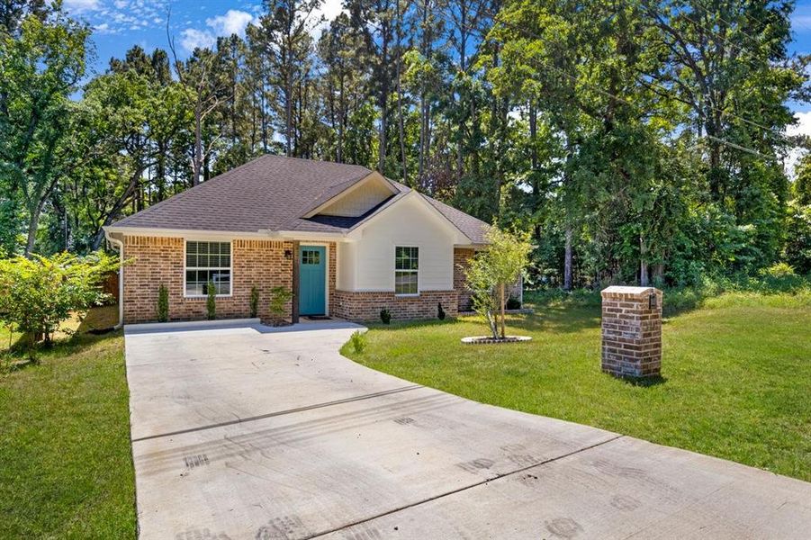 Single story home featuring concrete driveway, brick siding, roof with shingles, a front yard, and view of wooded area Single story home featuring concrete driveway, brick siding, roof with shingles, a front yard, and view of wooded area