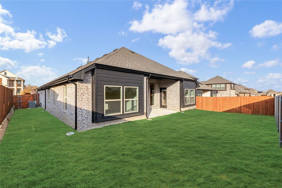 Back of single-story brick home with dark siding, large windows, patio, and fenced green backyard