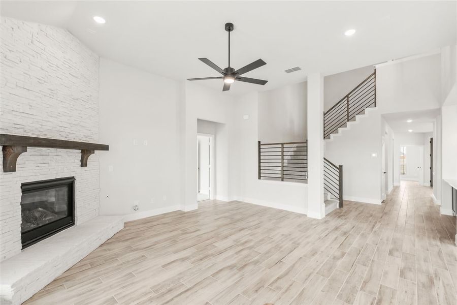 Open living room with stone fireplace, ceiling fan, light wood floors, and staircase with metal railing Open living room with stone fireplace, ceiling fan, light wood floors, and staircase with metal railing