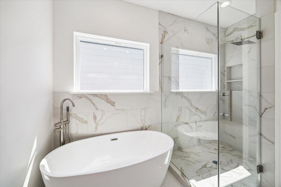 The primary bath’s freestanding soaking tub and oversized glass-enclosed shower create a luxurious focal point. Marble-look tile surrounds the bathing area and continues into the shower, where a rainfall-style showerhead and built-in niche enhance the modern design. Natural light from the windows brightens the space, while brushed metal fixtures add a refined finishing touch.