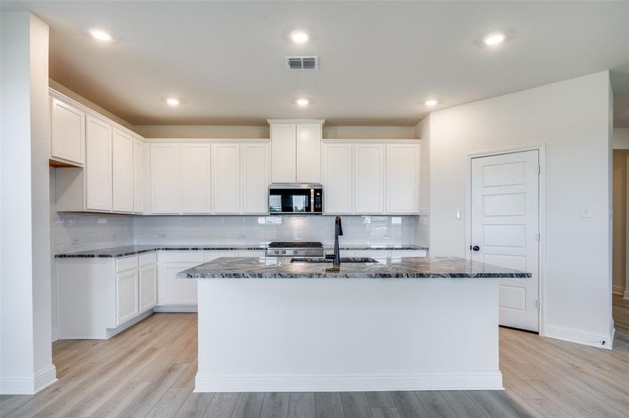Kitchen featuring dark stone countertops, a kitchen island with sink, stainless steel microwave, decorative backsplash, and recessed lighting Kitchen featuring dark stone countertops, a kitchen island with sink, stainless steel microwave, decorative backsplash, and recessed lighting
