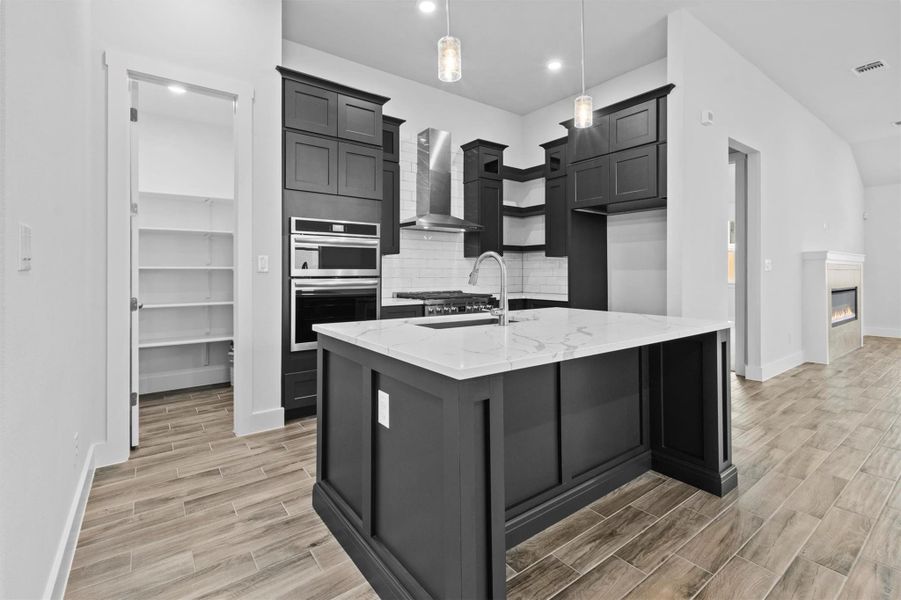 Kitchen featuring a center island with sink, stainless steel double oven, pendant lighting, light stone countertops, and wall chimney exhaust hood