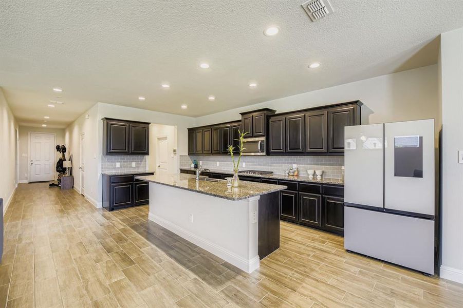 Kitchen featuring a textured ceiling, freestanding refrigerator, light stone countertops, recessed lighting, and a kitchen island with sink Kitchen featuring a textured ceiling, freestanding refrigerator, light stone countertops, recessed lighting, and a kitchen island with sink