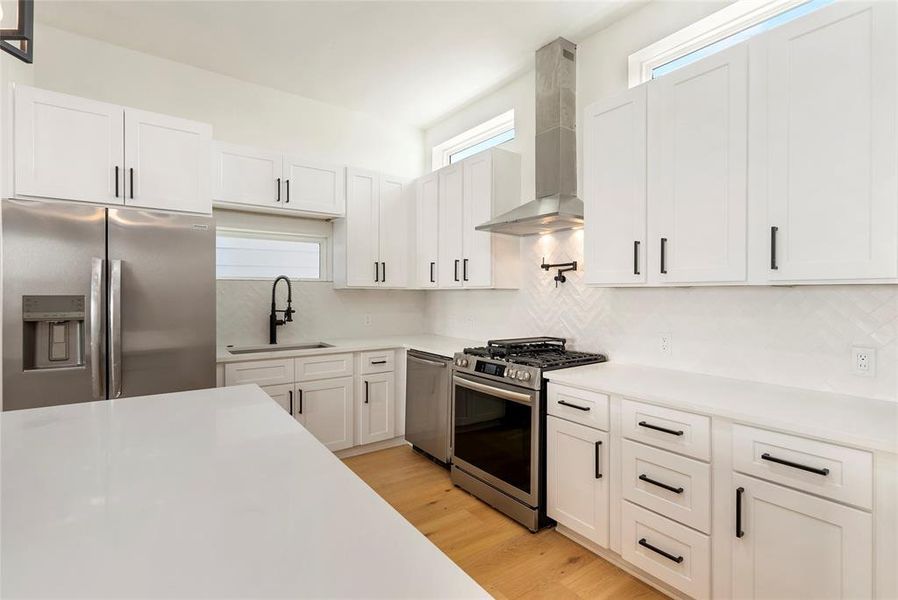 This is a modern kitchen featuring white cabinetry, stainless steel appliances, a gas stove with pot filler,  and a central island with a white countertop. It has a clean, contemporary design with a herringbone tile backsplash.