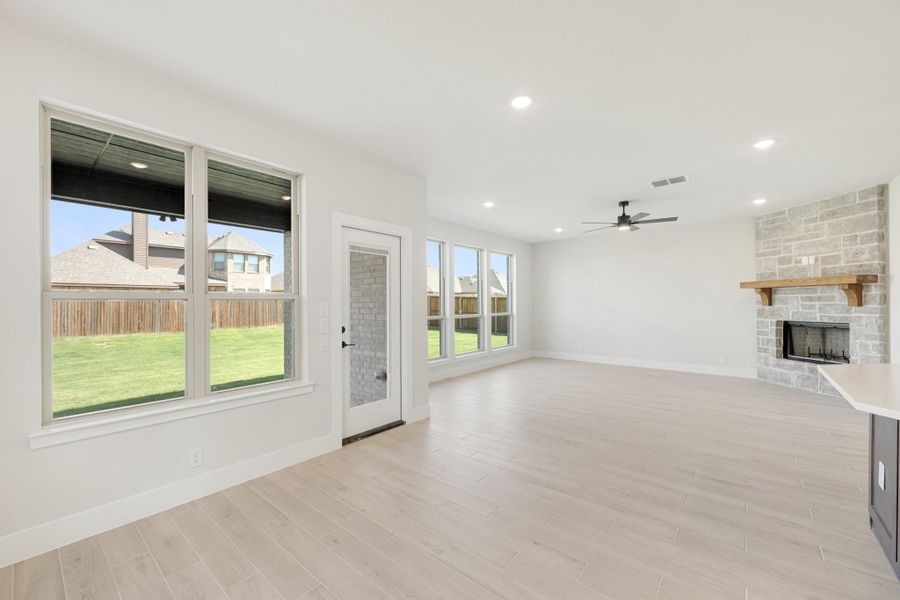 Open living room with light wood floors, stone fireplace, ceiling fan, and large windows overlooking backyard
