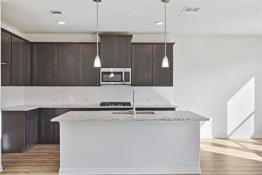 Kitchen with dark brown cabinets, light hardwood / wood-style floors, an island with sink, light stone counters, and backsplash