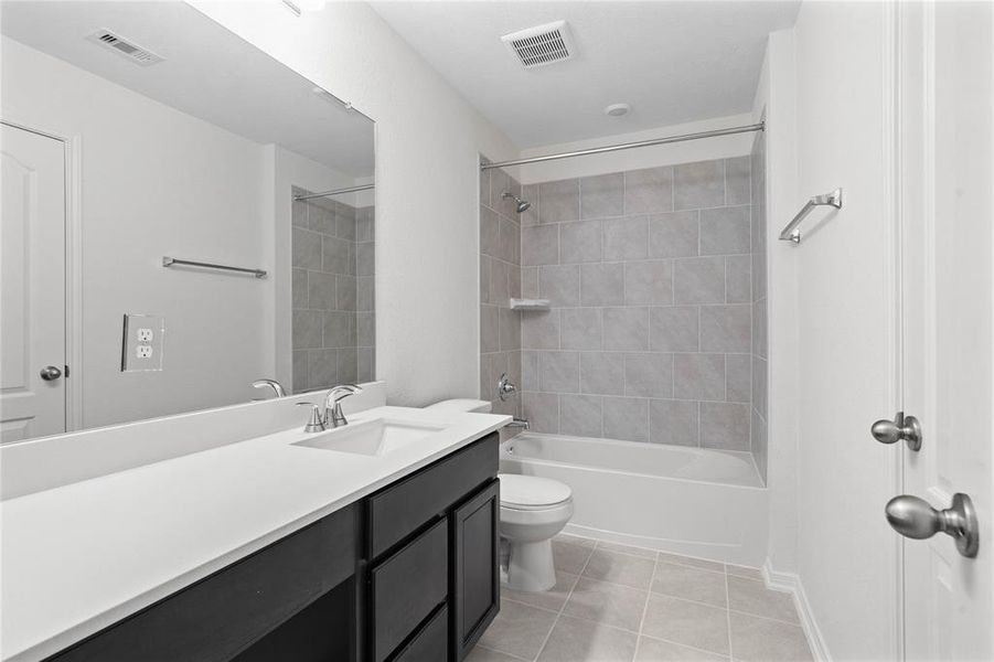 Secondary bath features tile flooring, bath/shower combo with tile surround, stained wood cabinets, beautiful light countertops, mirror, dark, sleek fixtures and modern finishes.