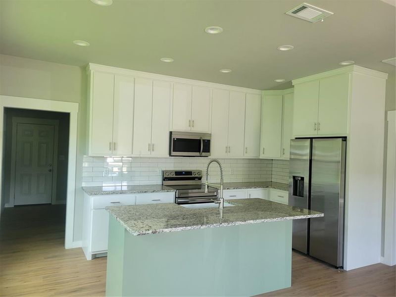 Modern kitchen featuring an island with a sink, stainless steel appliances, white cabinetry, granite countertops, and subway tile backsplash