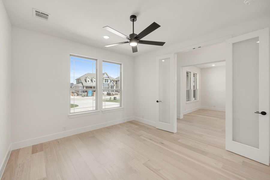 Empty bedroom with light hardwood floors, ceiling fan, and two windows overlooking a neighborhood