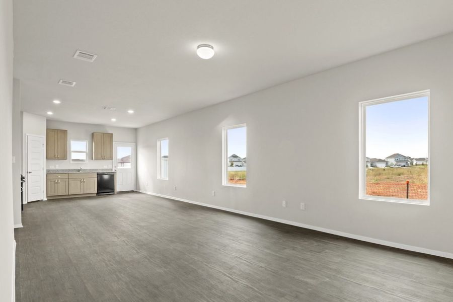 Image of a living room with brown flooring and light grey walls with windows and kitchen in the distance