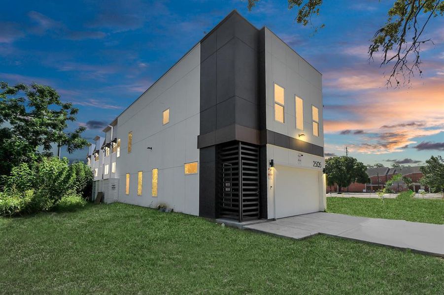 This is a modern two-story home featuring a geometric exterior with contrasting dark and light panels, large windows for ample natural light, a spacious garage, and a green lawn, all captured against a beautiful evening sky.