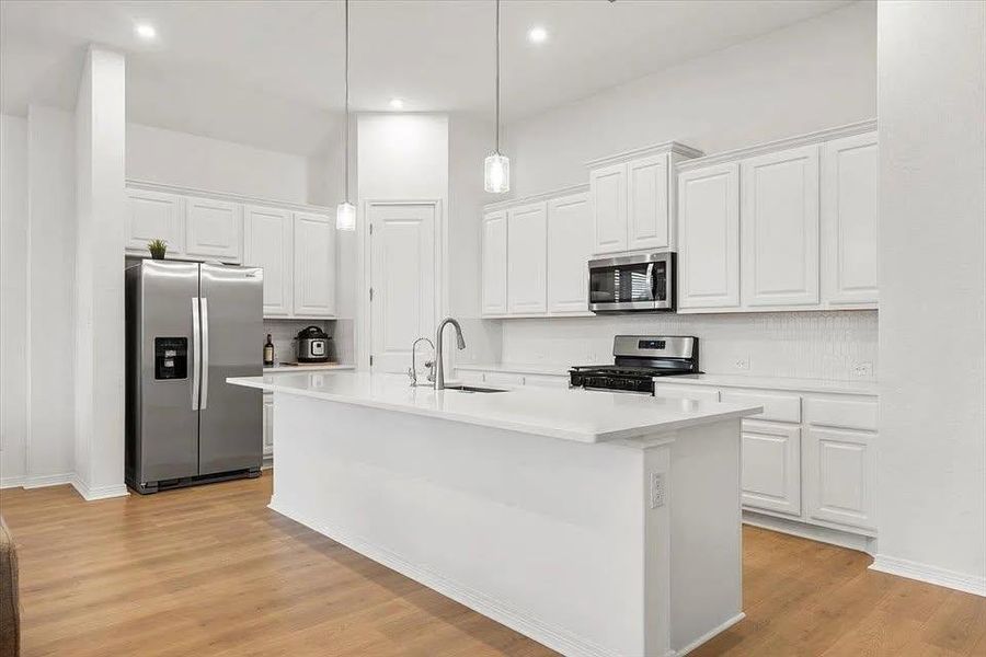 Kitchen featuring stainless steel appliances, white cabinets, light wood-style flooring, a center island with sink, and pendant lighting Kitchen featuring stainless steel appliances, white cabinets, light wood-style flooring, a center island with sink, and pendant lighting