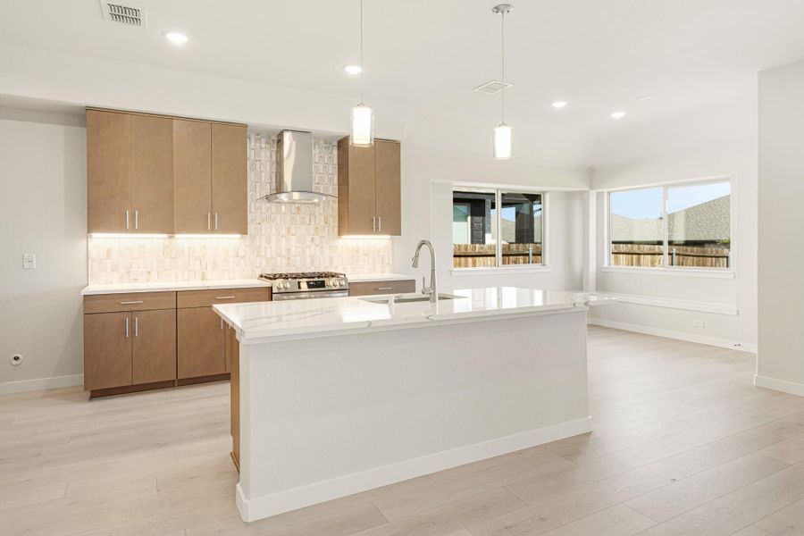 Open kitchen with large white island, brown cabinets, stainless range hood, and light wood floors