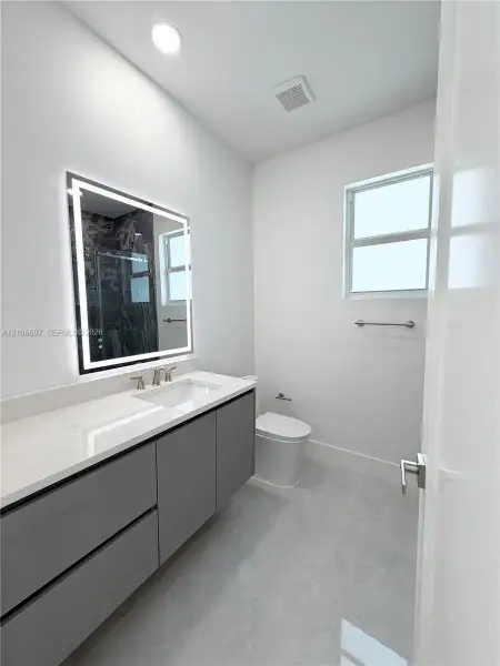 Modern vanity featuring a stone countertop, integrated sink, and illuminated rectangular mirror, complemented by grey cabinetry, light-toned flooring, and a window for natural light