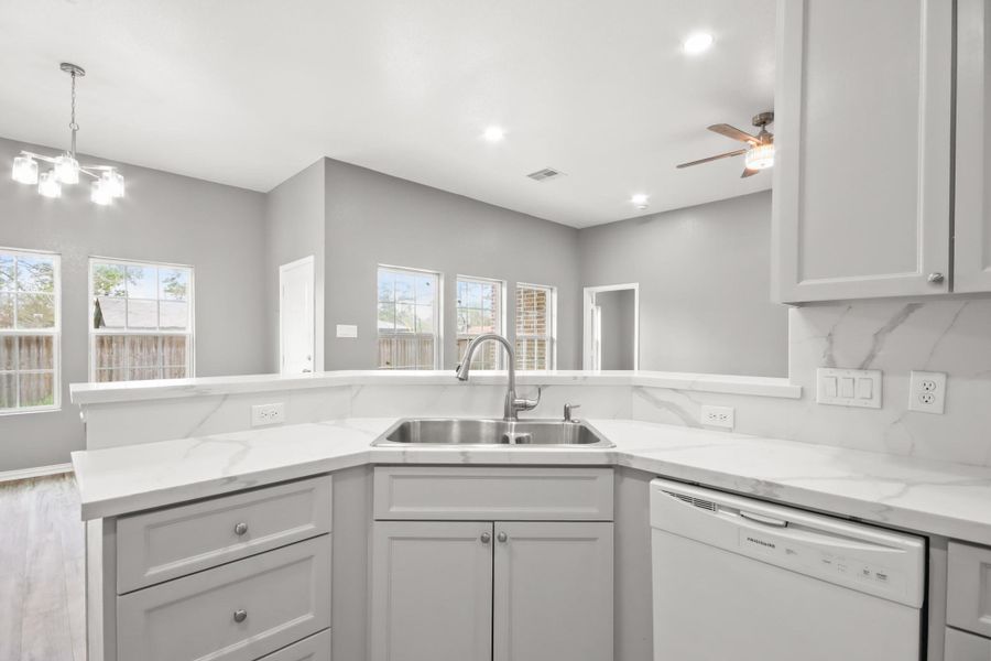 This modern kitchen features sleek gray cabinetry, marble-patterned countertops, and a stainless steel sink. It opens to a bright living area with large windows and a ceiling fan, offering a spacious and airy feel.