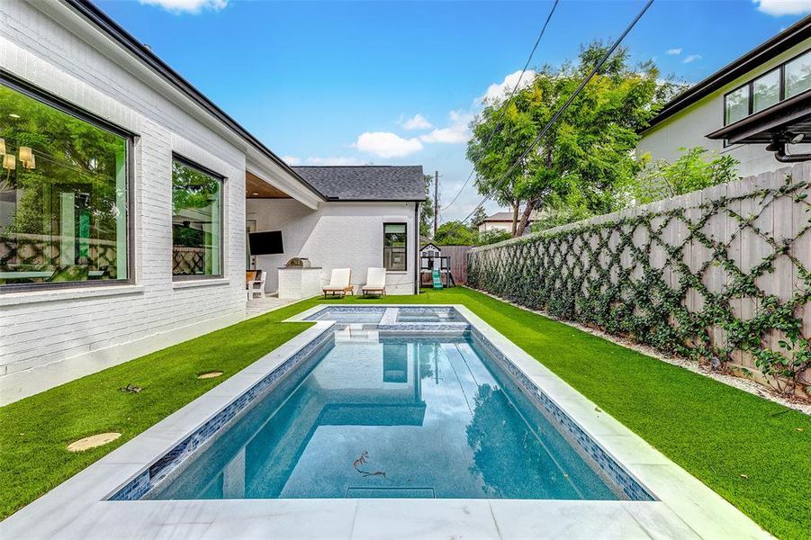 This photo showcases a modern backyard featuring a sleek rectangular pool with a spa, surrounded by lush artificial grass. The area includes a cozy patio with seating and a wall-mounted TV, perfect for outdoor relaxation and entertainment. The backyard also features a children's playhouse.