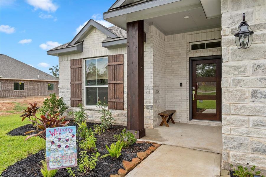 This welcoming entryway to the home featuring a clean white brick exterior, a wood front door with glass panels, and a charming porch area. The house has a pitched roof and a well-maintained garden bed conveying a warm, inviting atmosphere.