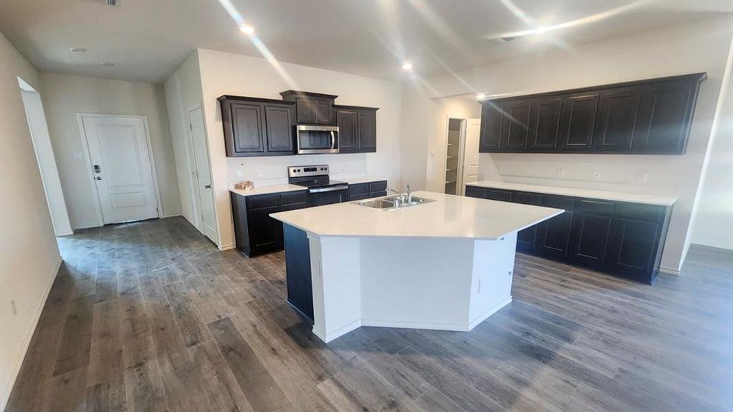Open-concept kitchen featuring an angular island with a sink, dark wood-finish cabinetry, stainless steel appliances, white countertops, and a white subway tile backsplash