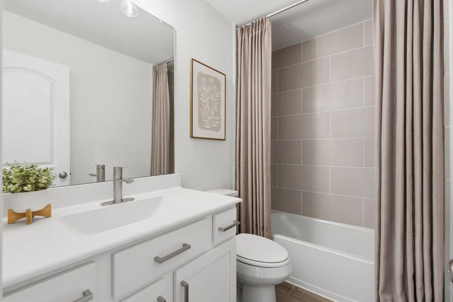 This bathroom features a modern design with a sleek white vanity, large mirror, and neutral-toned tiles. It includes a bathtub with a shower curtain and stylish fixtures, creating a clean and inviting space.