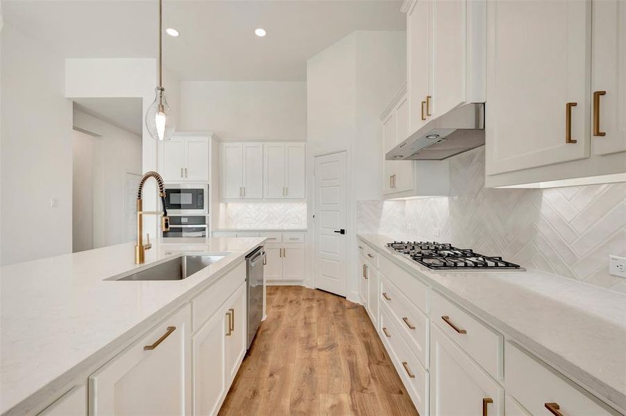 Modern kitchen featuring white cabinetry, light countertops, gold-tone hardware, wood-finish flooring, and a herringbone pattern tile backsplash