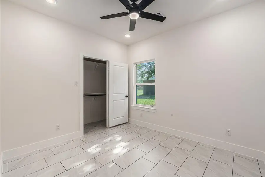 Bright room with large floor tiles, recessed lighting, and a ceiling fan. Features a window with a view of greenery and an open closet.