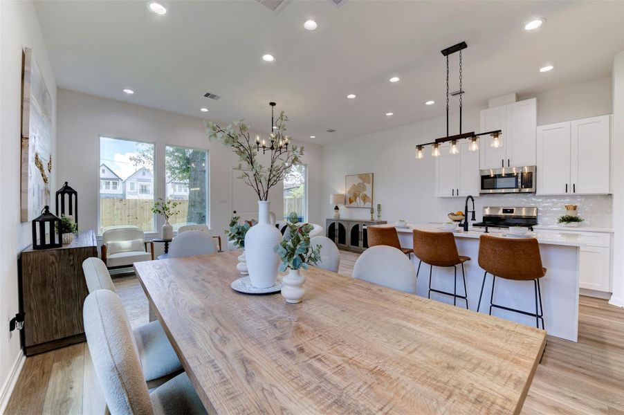View from dining table toward kitchen showing the full open-concept flow, hardwood floors, recessed lighting, dual matte black pendant chandeliers, island with three leather bar stools, white shaker cabinets, stainless appliances, and large rear-facing windows with backyard and street views. Living room sitting area visible in background.