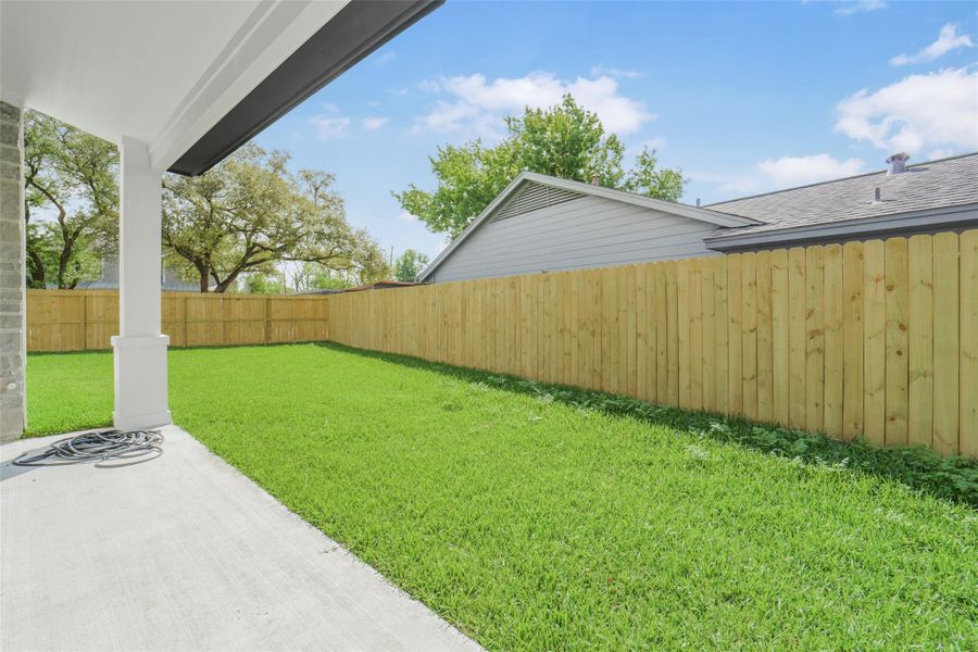 This photo showcases a spacious backyard with a well-maintained lawn, bordered by a wooden privacy fence. The area features a covered patio, ideal for outdoor relaxation, and is surrounded by mature trees, providing a serene atmosphere.