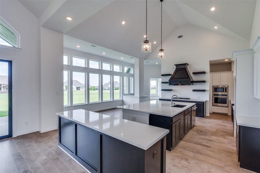 Kitchen featuring custom range hood, stainless steel appliances, an island with sink, light wood-type flooring, and high vaulted ceiling