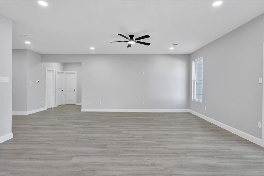 This photo showcases a spacious, modern living area with light gray walls, sleek wooden flooring, recessed lighting, and a ceiling fan. The room has ample natural light from a window, and there are multiple doors leading to other areas of the home, offering a clean, open feel.