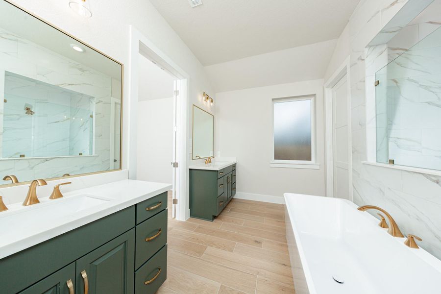 This spa-inspired primary bathroom showcases a sleek freestanding soaking tub set against a striking tiled accent wall with a glass-enclosed shower beyond. Tile flooring, clean modern lines, and brushed gold fixtures create a serene and contemporary retreat filled with natural light.