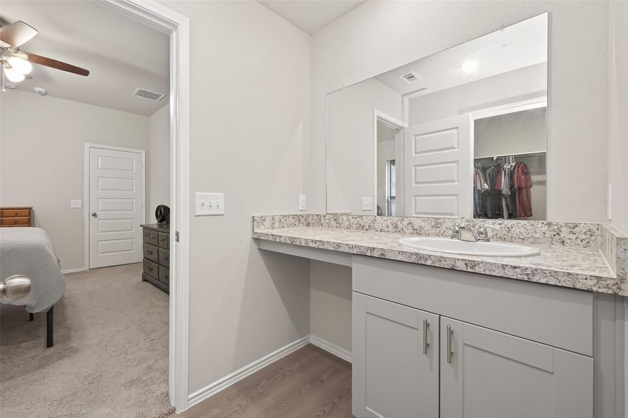 This photo features a modern bathroom with a spacious vanity, granite countertop, and large mirror. It connects to a carpeted bedroom with a ceiling fan and visible closet space.