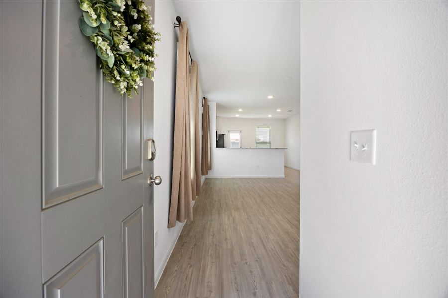 This photo shows a welcoming entryway with a light-colored door adorned with a wreath. It opens into a bright, open-concept living space featuring wood flooring, neutral walls, and modern lighting.