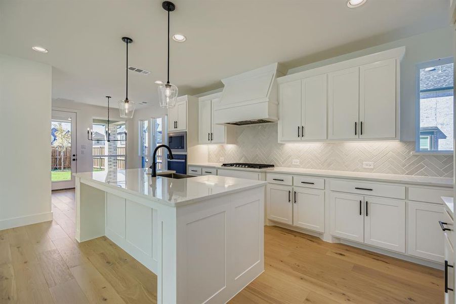 Kitchen featuring an island with sink, recessed lighting, hanging light fixtures, custom range hood, and white cabinetry Kitchen featuring an island with sink, recessed lighting, hanging light fixtures, custom range hood, and white cabinetry